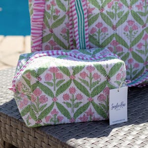 May include: A white floral print cosmetic bag and tote bag with pink and green accents. The bags feature a repeating pattern of pink flowers and green leaves. The tote bag has pink and green striped handles. A white tag with the text "Saylor Rae" is attached to the cosmetic bag.