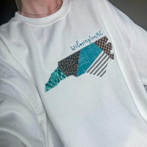 Puede incluir: Sudadera blanca con un diseño de patchwork colorido de la costa de Carolina del Norte. El diseño incluye patrones turquesa, negros y marrones, con el texto "Wilmington NC" en cursiva. La sudadera es de estilo casual y cómodo.