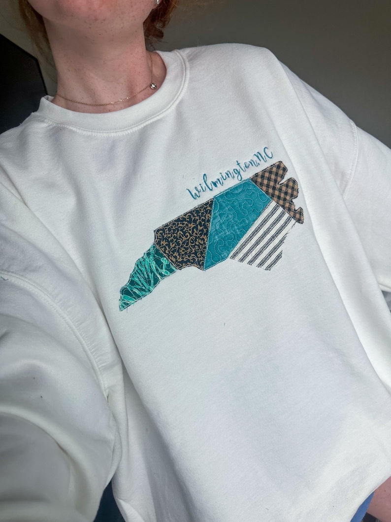 May include: White sweatshirt featuring a colorful, patchwork design of the North Carolina coastline. The design includes teal, black, and brown patterns, with the text "Wilmington NC" in cursive. The sweatshirt is a casual, comfortable style.