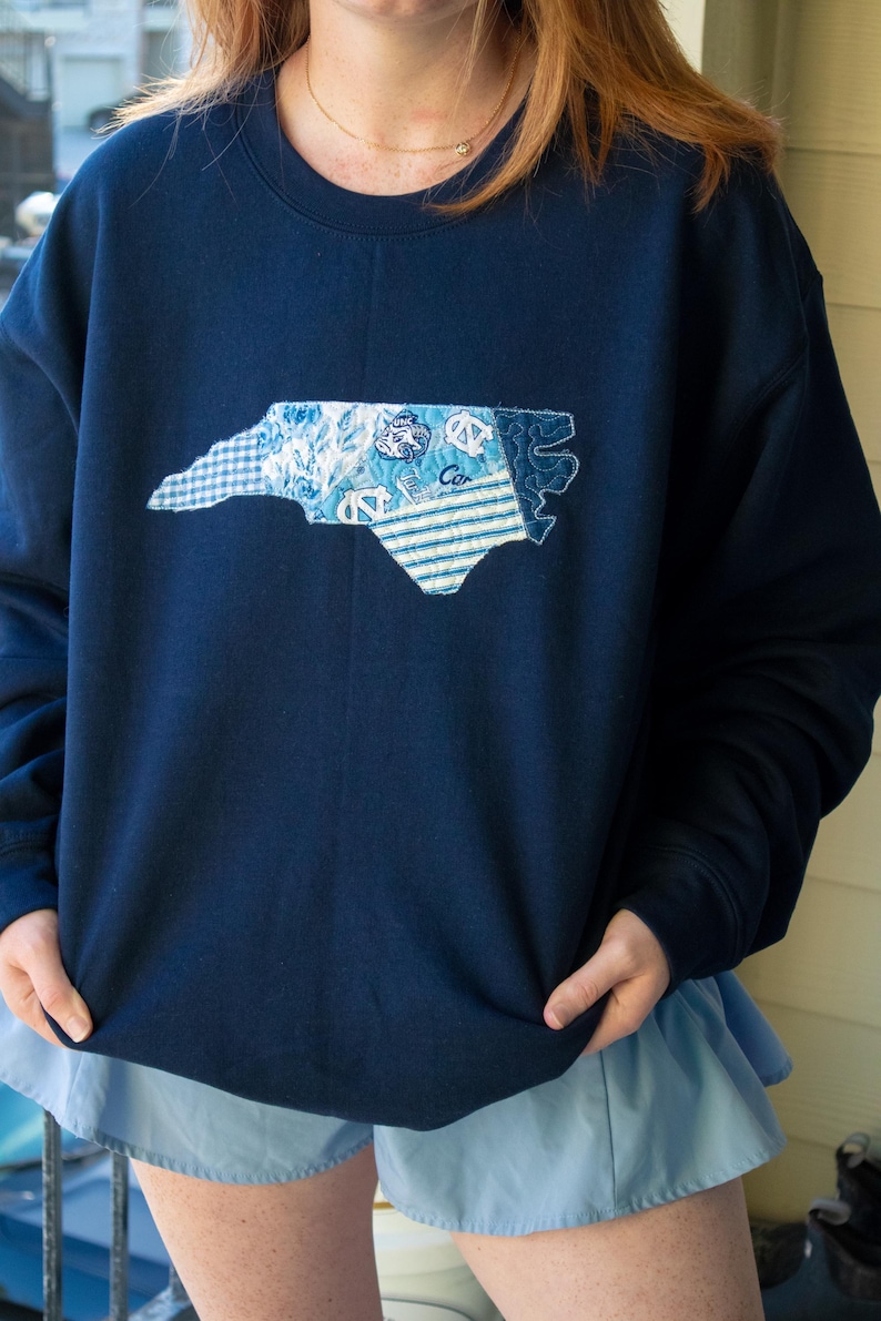 K&ouml;nnte beinhalten: Marineblaues Sweatshirt mit einem Patchwork-Design des Umrisses des Bundesstaates North Carolina. Der Patch enth&auml;lt verschiedene blau-wei&szlig;e Muster, darunter Streifen und Blumendrucke. Das Sweatshirt hat eine marineblaue Farbe.
