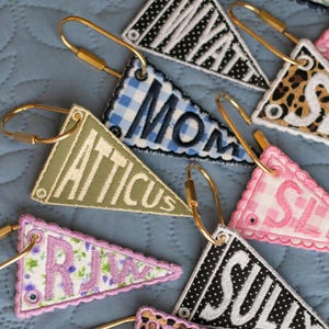 May include: Embroidered pennant keyrings in diverse patterns and colours, including gingham, floral, and leopard print. Each keyring features a name or word in a contrasting colour, attached to a gold-tone clasp.