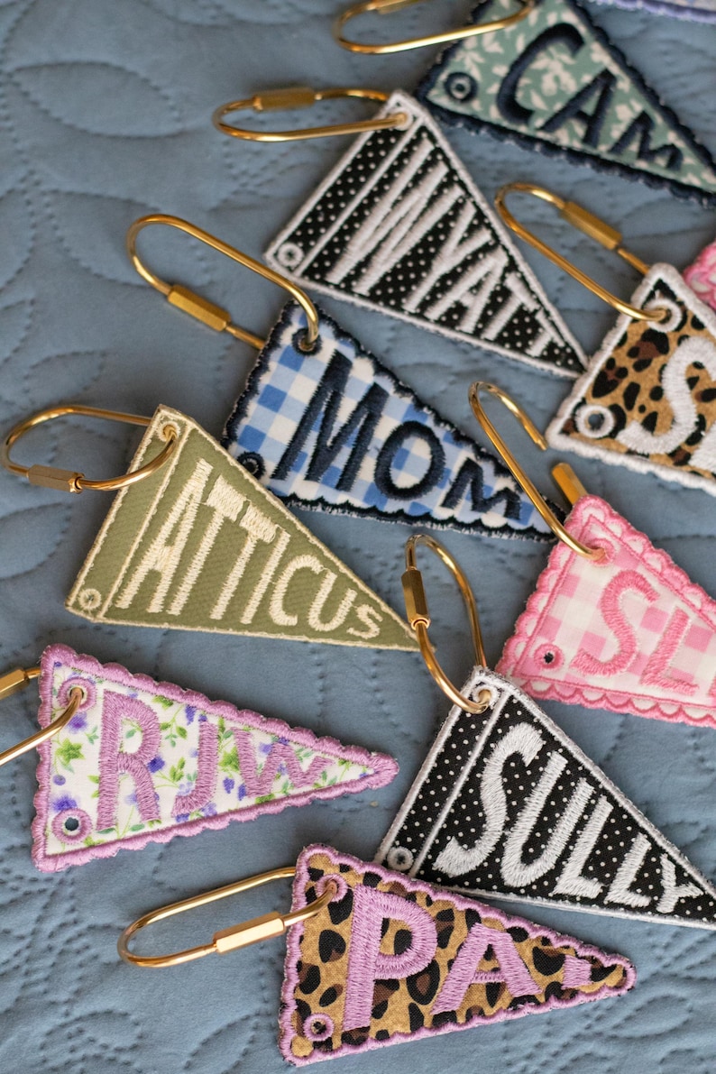May include: Embroidered pennant keyrings in diverse patterns and colours, including gingham, floral, and leopard print. Each keyring features a name or word in a contrasting colour, attached to a gold-tone clasp.