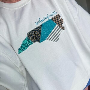 May include: White sweatshirt featuring a colorful, patchwork design of the North Carolina coastline. The design includes teal, navy, and brown patterned fabrics, with the word "Wilmington nc" in script above the design. The sweatshirt has a crew neck.