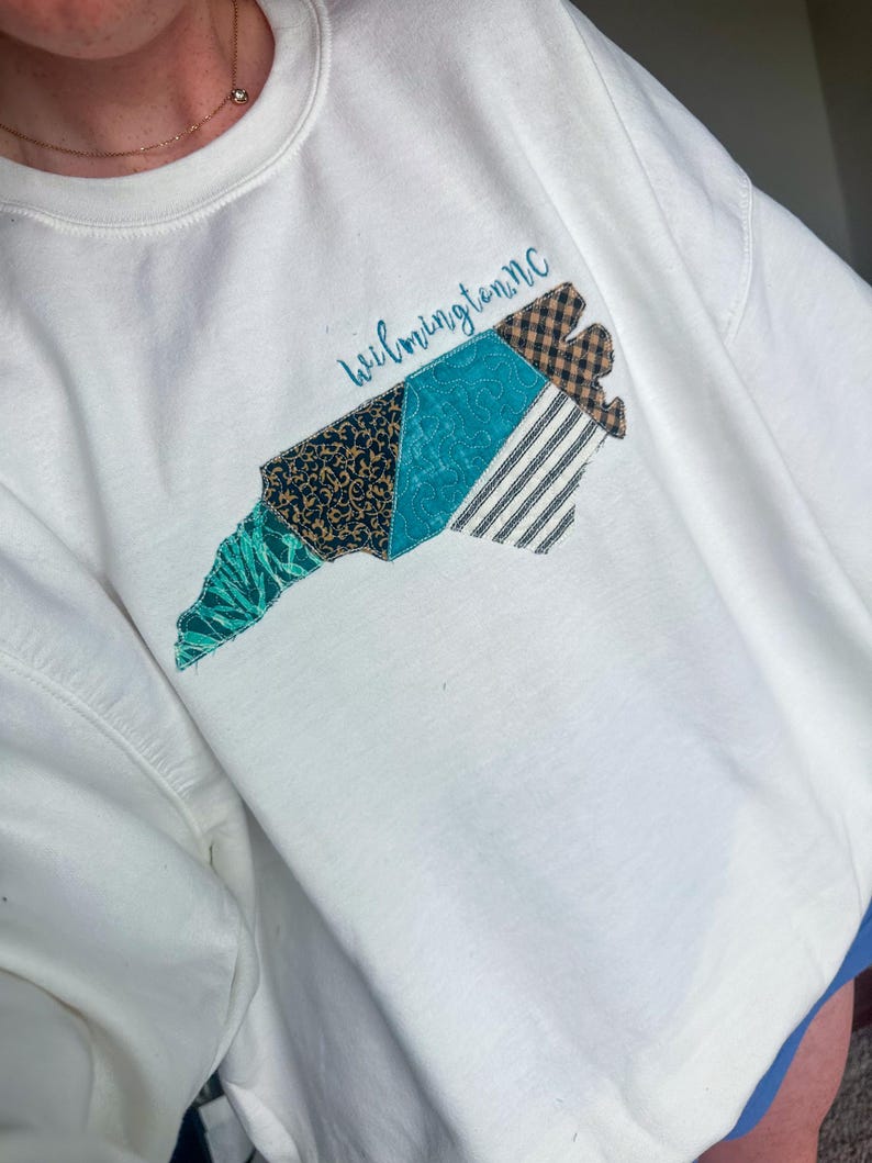 May include: White sweatshirt featuring a colorful, patchwork design of the North Carolina coastline. The design includes teal, navy, and brown patterned fabrics, with the word "Wilmington nc" in script above the design. The sweatshirt has a crew neck.