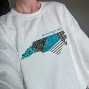 May include: White sweatshirt featuring a patchwork design of the North Carolina coastline. The design incorporates various patterns and colors, including teal, black, and brown. The text "Wilmington, NC" is written in cursive above the design.