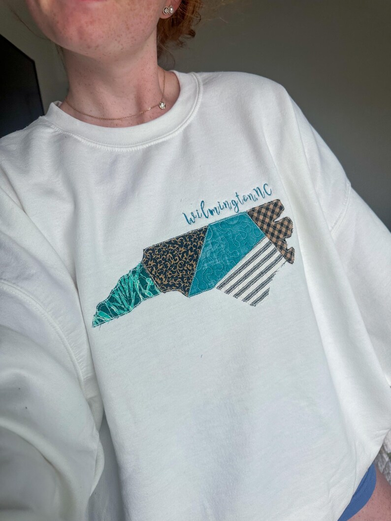 May include: White sweatshirt featuring a patchwork design of the North Carolina coastline. The design incorporates various patterns and colors, including teal, black, and brown. The text "Wilmington, NC" is written in cursive above the design.