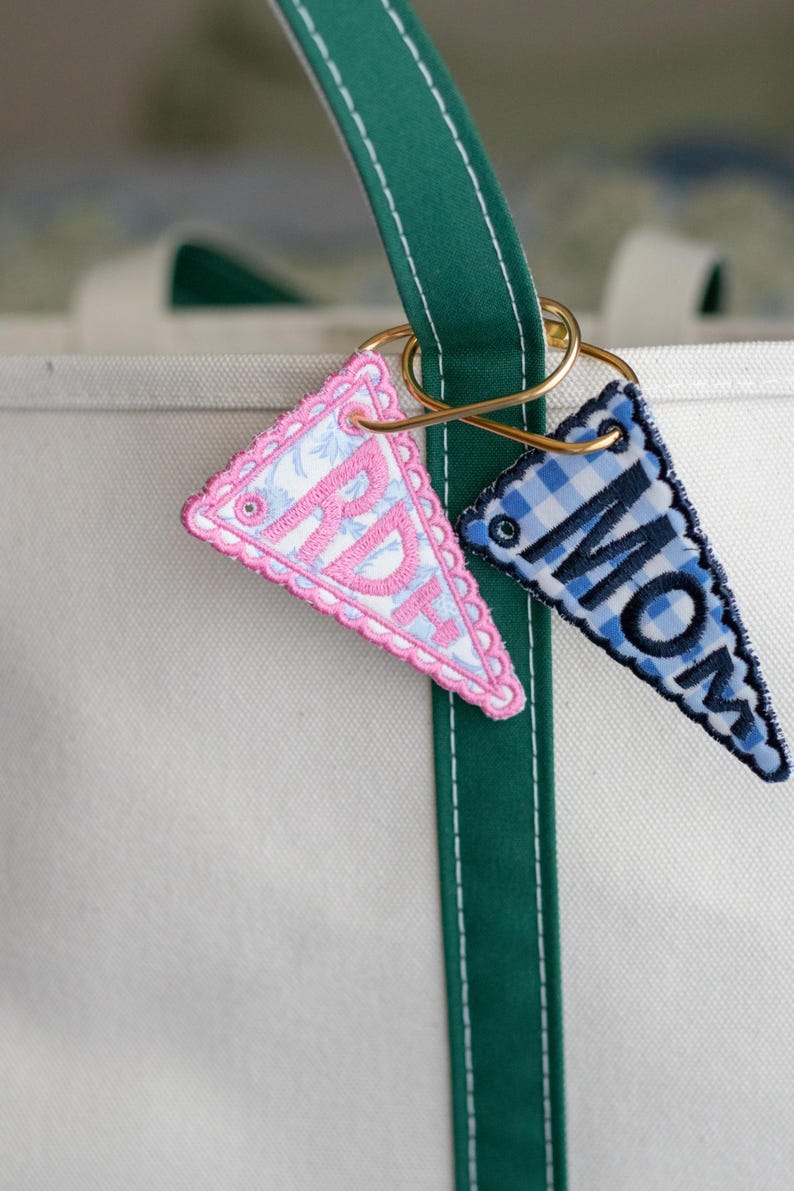 May include: Two embroidered pennant keyrings hang from a gold ring on a green and white tote bag. One pink pennant has the letters "RD" and the other blue pennant has the word "MOM". The pennants have a scalloped edge.