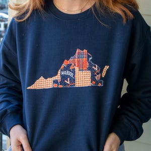 May include: Navy blue sweatshirt featuring a patchwork design of the state of Virginia. The design incorporates orange, blue, and white plaid and gingham patterns. The word "WAHOOS" is printed on the design.