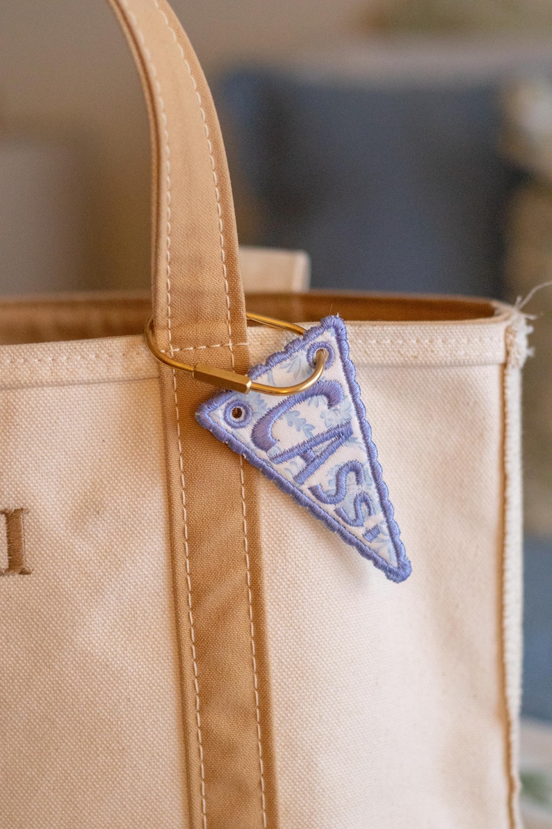 May include: A beige canvas tote bag with brown straps and a gold-coloured metal ring holding a small, triangular pennant. The pennant is light blue with the name "CASS" embroidered in a darker blue font.