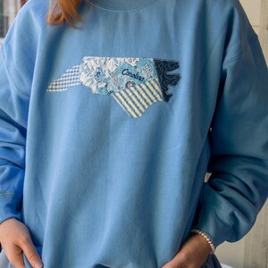 Op de afbeelding: Lichtblauw sweatshirt met een applicatie-omtrek van North Carolina. De applicatie bevat verschillende blauwe stoffen met patronen, waaronder bloemen- en streepontwerpen, met het woord "Carolina" erop gestikt. De sweatshirt heeft lange mouwen.