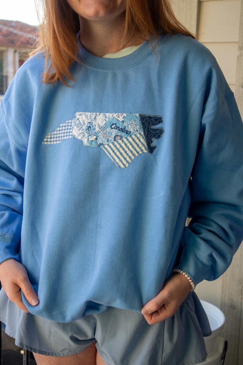 Op de afbeelding: Lichtblauw sweatshirt met een applicatie-omtrek van North Carolina. De applicatie bevat verschillende blauwe stoffen met patronen, waaronder bloemen- en streepontwerpen, met het woord "Carolina" erop gestikt. De sweatshirt heeft lange mouwen.