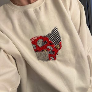 Puede incluir: Sudadera blanca con un diseño de patchwork rojo, blanco y negro del estado de Ohio. El diseño incluye la palabra "Buckeyes" y un casco de fútbol.