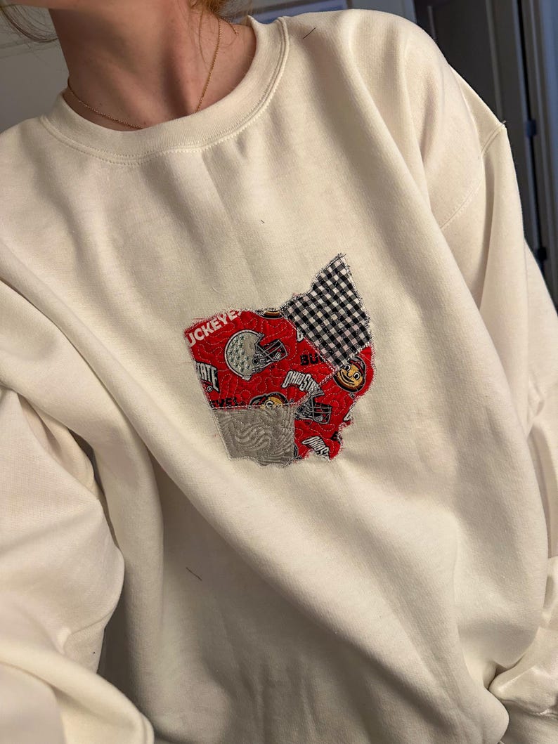 Op de afbeelding: Witte trui met een patchwork-ontwerp in rood, wit en zwart van de staat Ohio. Het ontwerp bevat het woord "Buckeyes" en een voetbalhelm.