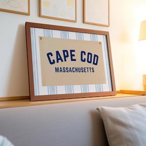 Op de afbeelding: Ingelijste wandkunst met de tekst "CAPE COD MASSACHUSETTS" in marineblauw op een beige achtergrond. Het kunstwerk is ingelijst in een bruine houten lijst en heeft een lichtblauwe en witte gestreepte rand. De kunst wordt op een plank tentoongesteld.