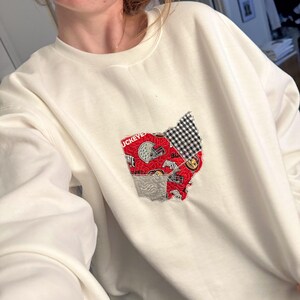 Op de afbeelding: Witte trui met een rood en zwart patchwork ontwerp van de staat Ohio. Het ontwerp bevat het woord "Buckeye" en een smiley.