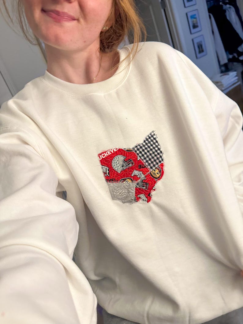 Op de afbeelding: Witte trui met een rood en zwart patchwork ontwerp van de staat Ohio. Het ontwerp bevat het woord "Buckeye" en een smiley.