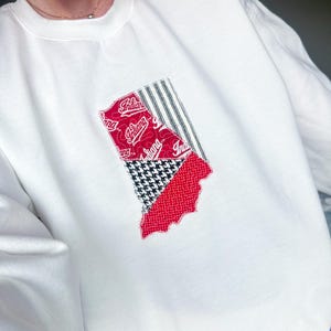 May include: White sweatshirt with an Indiana-shaped patch. The patch incorporates red, black, and white patterns, including houndstooth and striped designs, and the word "Indiana".