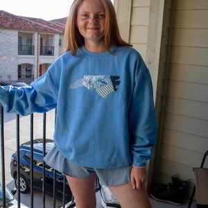 Op de afbeelding: Een lichtblauw sweatshirt met een patchwork ontwerp van de omtrek van de staat North Carolina. Het patchwork bevat bloemen-, gestreepte en effen blauwe stoffen. Het woord "Carolina" is zichtbaar. Het sweatshirt wordt gedragen met een blauwe short.