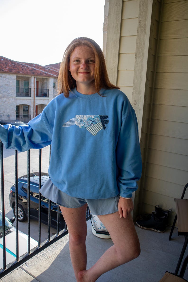 Op de afbeelding: Een lichtblauw sweatshirt met een patchwork ontwerp van de omtrek van de staat North Carolina. Het patchwork bevat bloemen-, gestreepte en effen blauwe stoffen. Het woord "Carolina" is zichtbaar. Het sweatshirt wordt gedragen met een blauwe short.