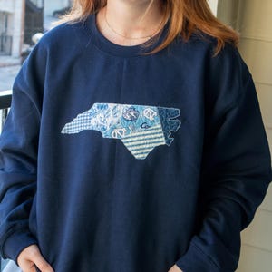 K&ouml;nnte beinhalten: Marineblaues Sweatshirt mit einem aufgen&auml;hten Umriss von North Carolina in verschiedenen blau gemusterten Stoffen. Das Sweatshirt mit Rundhalsausschnitt ist aus einem weichen Material gefertigt. Es handelt sich um ein Bekleidungsst&uuml;ck zum Thema Bundesstaat.