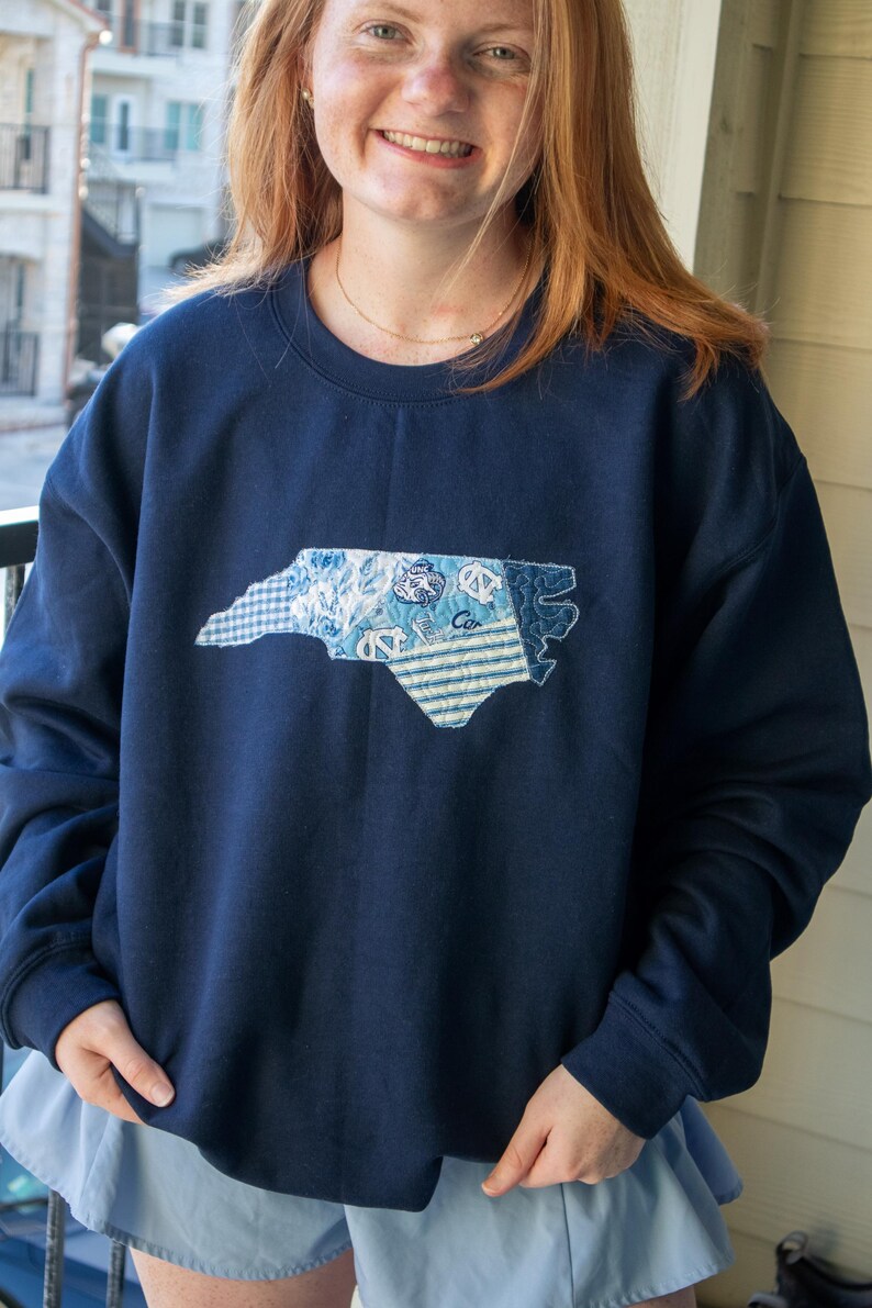 K&ouml;nnte beinhalten: Marineblaues Sweatshirt mit einem aufgen&auml;hten Umriss von North Carolina in verschiedenen blau gemusterten Stoffen. Das Sweatshirt mit Rundhalsausschnitt ist aus einem weichen Material gefertigt. Es handelt sich um ein Bekleidungsst&uuml;ck zum Thema Bundesstaat.