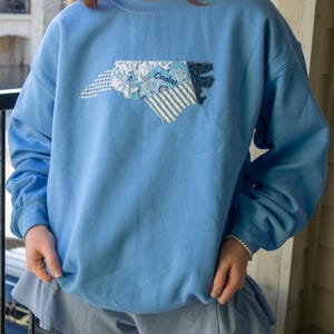 Op de afbeelding: Lichtblauw sweatshirt met een geborduurde omtrek van de staat North Carolina. De omtrek is gemaakt van verschillende stoffen met patronen, waaronder bloemen-, streep- en ruitmotieven. Het woord "Carolina" is op het ontwerp geborduurd.