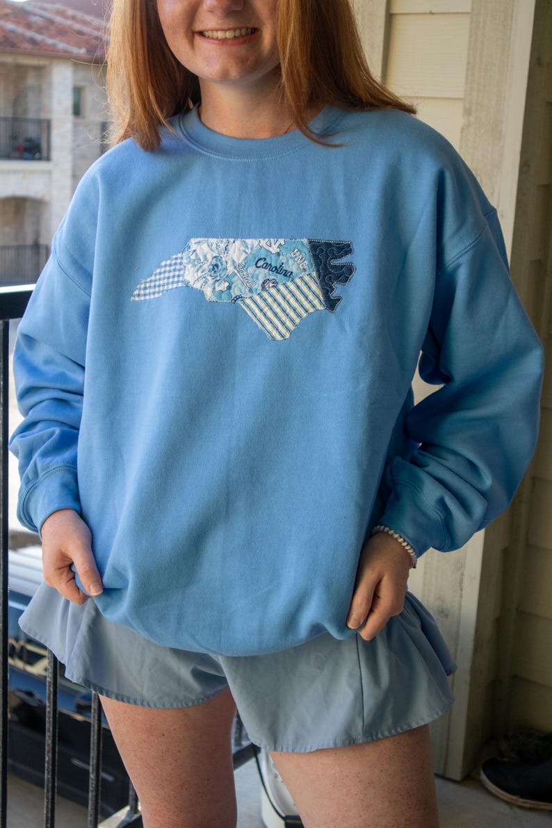 Op de afbeelding: Lichtblauw sweatshirt met een geborduurde omtrek van de staat North Carolina. De omtrek is gemaakt van verschillende stoffen met patronen, waaronder bloemen-, streep- en ruitmotieven. Het woord "Carolina" is op het ontwerp geborduurd.