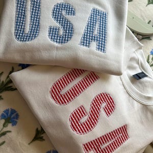 Op de afbeelding: Twee witte sweatshirts met het woord "USA" geborduurd. Het bovenste sweatshirt heeft blauw-wit geruite letters, en het onderste sweatshirt heeft rood-wit gestreepte letters.