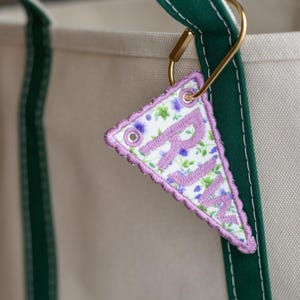 May include: A triangular fabric keyring embroidered with the letters "RJM" in lavender. The keyring features a floral pattern with purple and green accents, a scalloped edge, and is attached to a gold-coloured metal clasp.