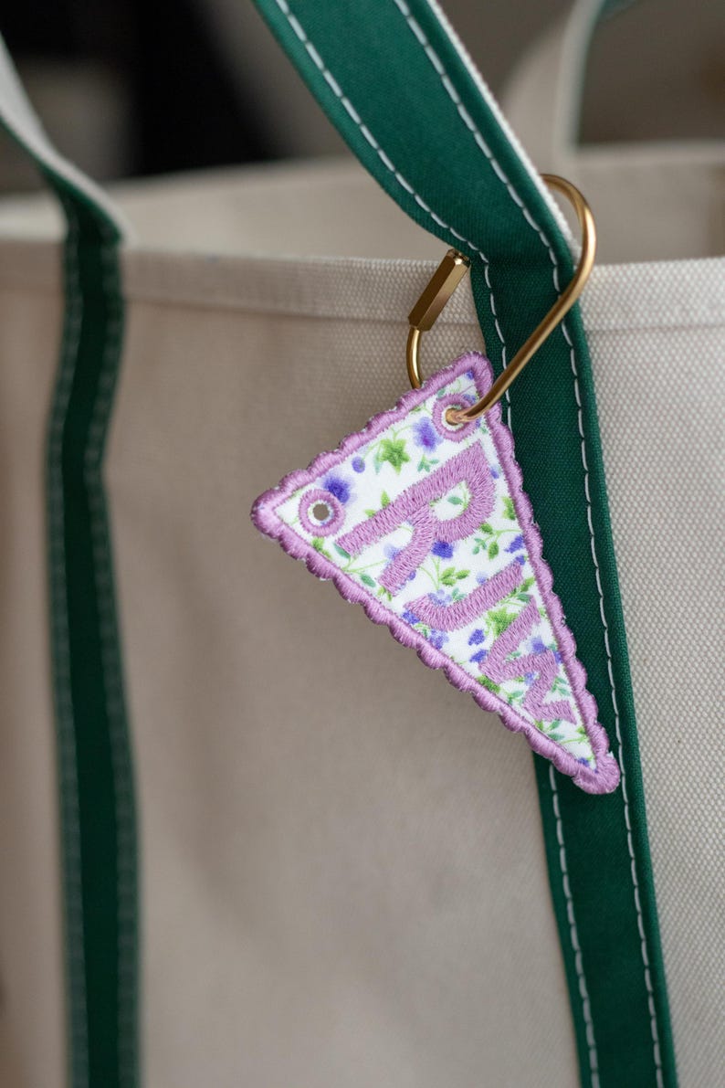 May include: A triangular fabric keyring embroidered with the letters "RJM" in lavender. The keyring features a floral pattern with purple and green accents, a scalloped edge, and is attached to a gold-coloured metal clasp.