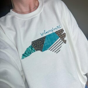 May include: White sweatshirt featuring a colorful patchwork design in the shape of North Carolina. The design includes teal, black, and brown patterned sections, with the text "Wilmington, NC" in a teal script. The sweatshirt has a crew neck and long sleeves.