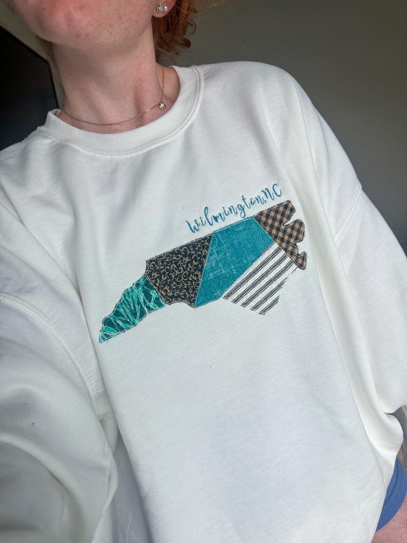 May include: White sweatshirt featuring a colorful patchwork design in the shape of North Carolina. The design includes teal, black, and brown patterned sections, with the text "Wilmington, NC" in a teal script. The sweatshirt has a crew neck and long sleeves.