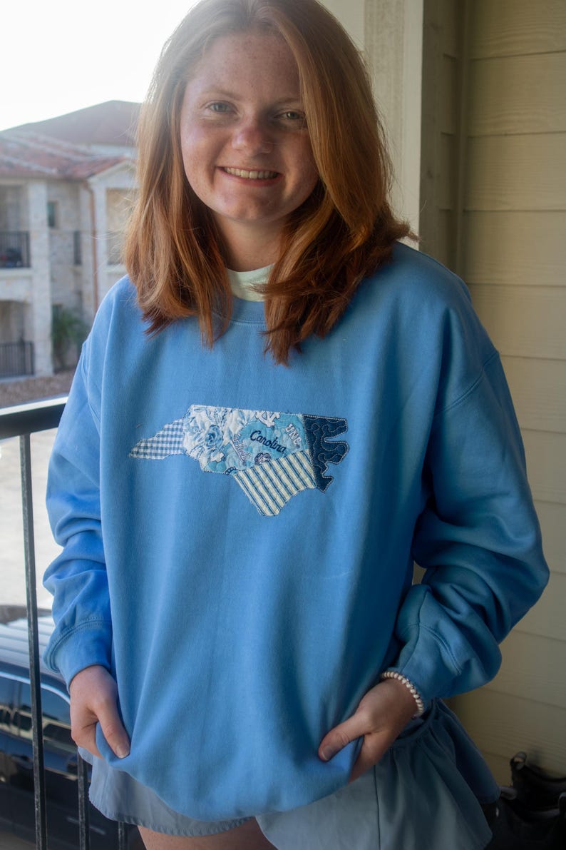 Op de afbeelding: Lichtblauw sweatshirt met een patch van de omtrek van de staat North Carolina. De patch is gemaakt van verschillende stoffen met patronen, waaronder bloemen- en streepontwerpen, met het woord "Carolina" erop gestikt.
