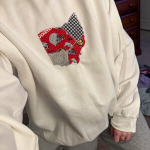 Op de afbeelding: Witte longsleeve sweatshirt met een rood, wit en blauw patchwork ontwerp van de staat Ohio. Het ontwerp bevat de woorden "Ohio State" en een voetbalhelm.