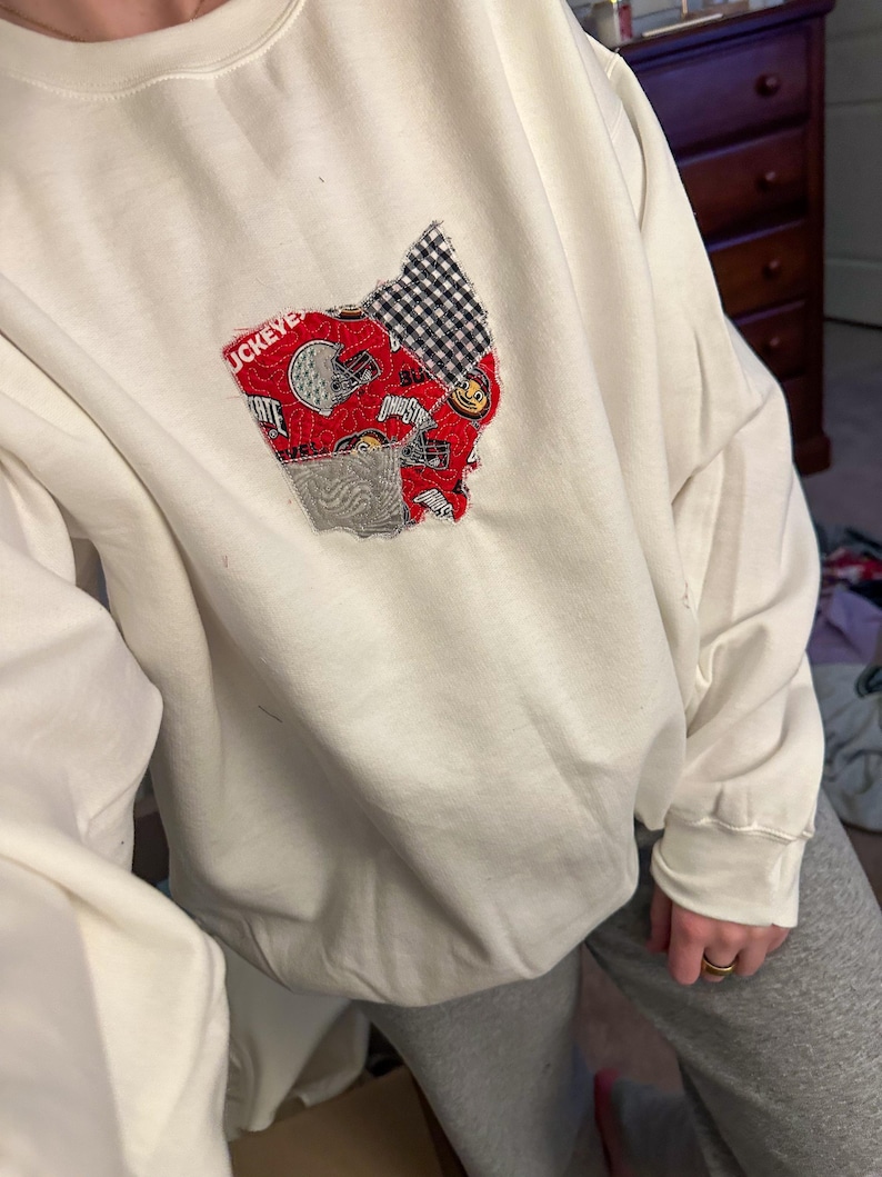 Op de afbeelding: Witte longsleeve sweatshirt met een rood, wit en blauw patchwork ontwerp van de staat Ohio. Het ontwerp bevat de woorden "Ohio State" en een voetbalhelm.