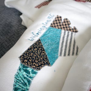 May include: White fabric item with a patchwork design in the shape of a state, featuring various patterns and colors. The word "Wilmington NC" is embroidered in blue. The patchwork includes teal, black, and brown patterns.