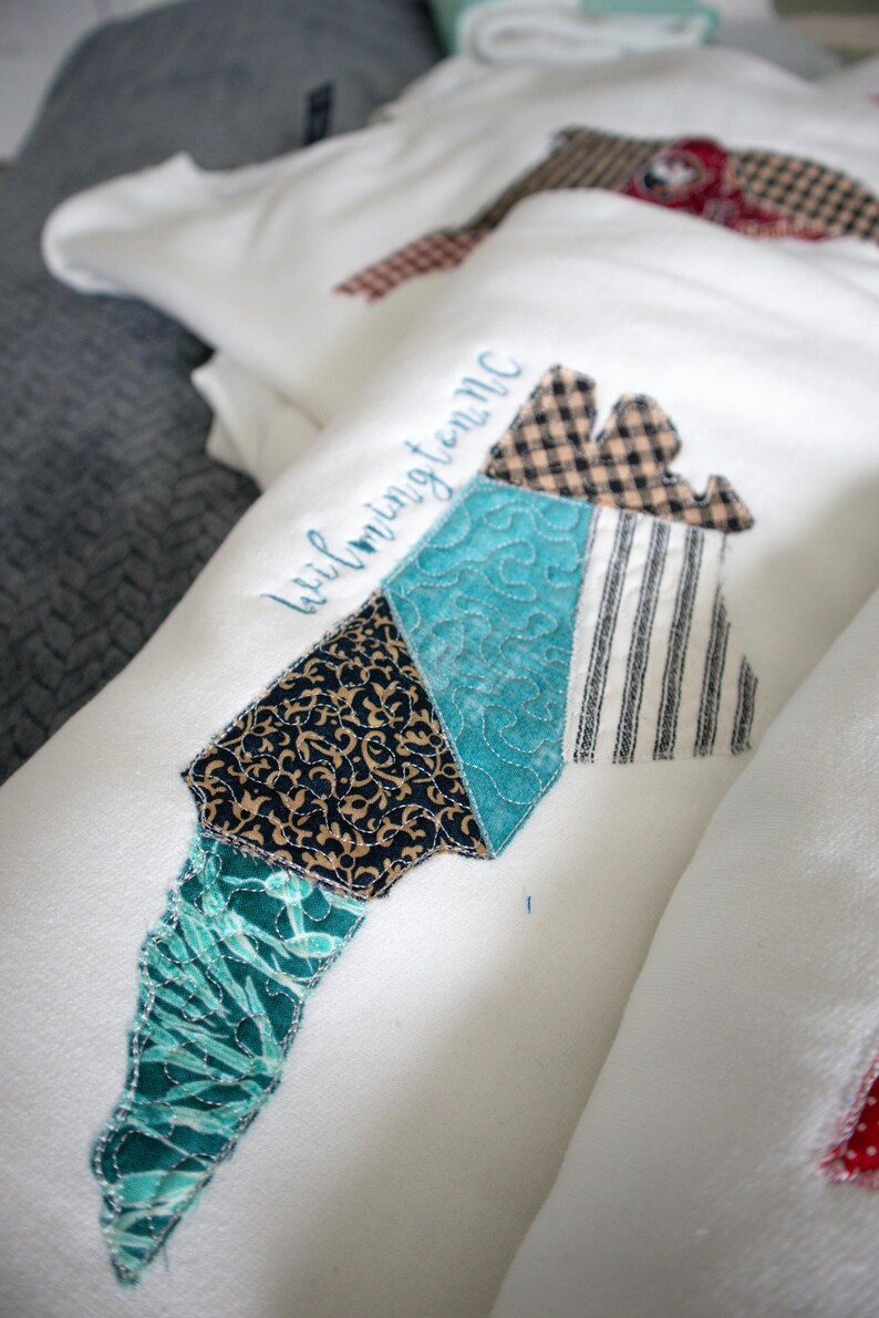 May include: White fabric item with a patchwork design in the shape of a state, featuring various patterns and colors. The word "Wilmington NC" is embroidered in blue. The patchwork includes teal, black, and brown patterns.