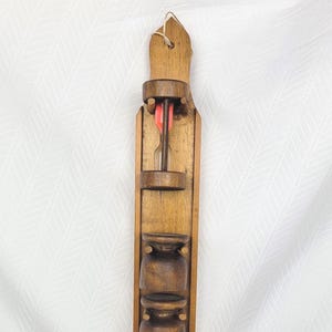 May include: A vintage wooden dart holder with a natural finish. The holder features carved cup-like compartments and a slot for the dart. A red dart is visible. The piece is designed to be hung on a wall.