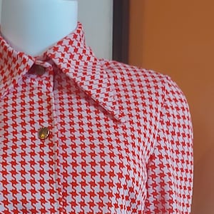 May include: A red and white houndstooth patterned shirt with a collared neckline and long sleeves. The shirt has a button-up front and a patch pocket on the left side.