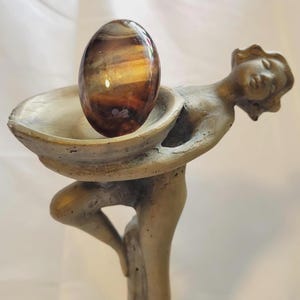 May include: A polished, oval-shaped gemstone with bands of brown, gold, and amber hues rests in a decorative, antique-style statue. The statue depicts a woman holding a shallow bowl. The gemstone is approximately 2 inches long.
