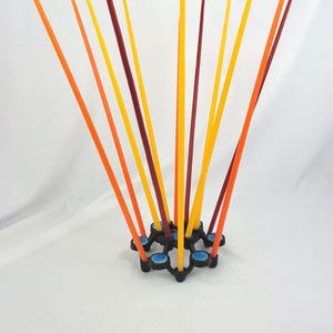May include: A black candle holder with a circular, multi-holed base featuring blue accents, holding a collection of tall, thin taper candles in shades of yellow, orange, and deep red, arranged in a radiating, fan-like pattern.