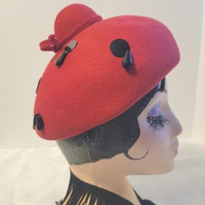 Peut inclure: Un chapeau rouge vif avec une petite couronne arrondie et un nœud décoratif. Le chapeau est orné de plusieurs ornements géométriques noirs. Le chapeau est présenté sur une tête de mannequin avec des cheveux noirs et une frange noire.