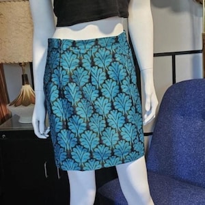 May include: A mannequin displays a black crop top and a turquoise mini-skirt with a leaf pattern. The skirt is the focal point, with its vibrant color and intricate design. The mannequin is on a patterned rug.