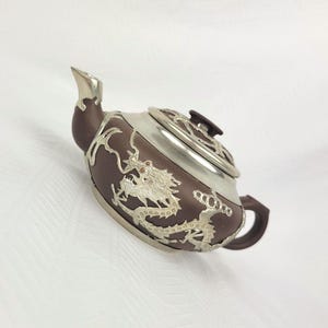 May include: A dark brown teapot featuring silver-coloured metal overlays. The body is decorated with an embossed silver dragon motif, while the lid, spout, and handle accents are crafted from a smooth, polished silver-toned metal material.