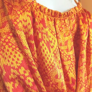 May include: Close-up of a patterned dress in shades of orange and yellow. The fabric has a repeating design and is gathered at the neckline. A yellow tie belt is visible at the waist. The dress is made of a lightweight material.