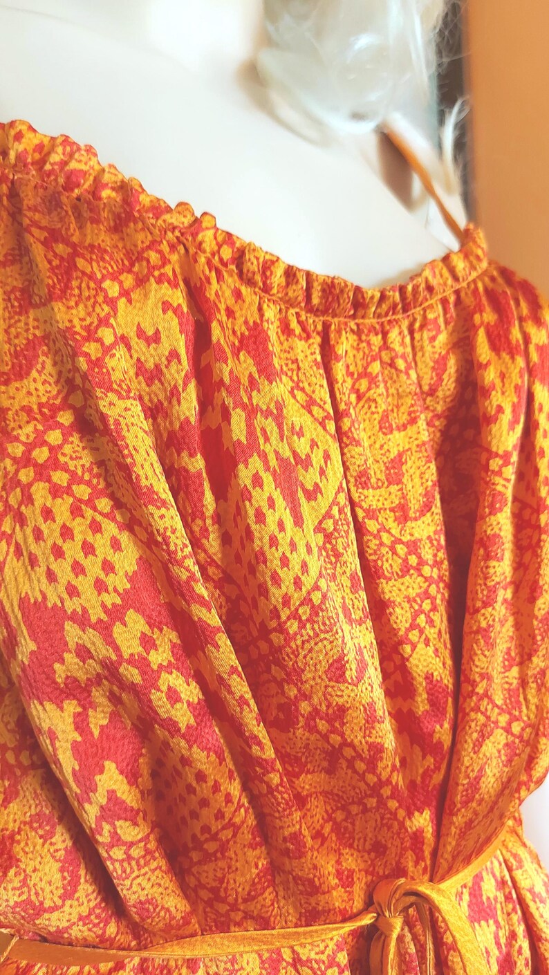 May include: Close-up of a patterned dress in shades of orange and yellow. The fabric has a repeating design and is gathered at the neckline. A yellow tie belt is visible at the waist. The dress is made of a lightweight material.