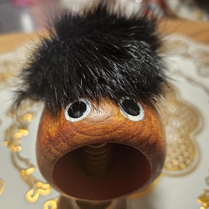 May include: A wooden nutcracker with a black fur hat and painted eyes. The nutcracker has a large open mouth and a brown wooden body.