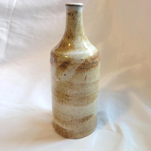 May include: A ceramic bottle with a narrow neck and a cylindrical body. The surface features a mottled glaze with earthy tones of beige, cream, and brown, displaying horizontal bands of colour and subtle speckling throughout the exterior finish.