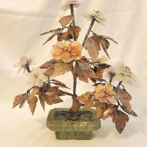 May include: A decorative bonsai tree sculpture with a dark brown trunk, bronze-colored leaves, and flowers in shades of white, peach, and yellow. The tree is set in a square, green stone pot. A unique home decor item.