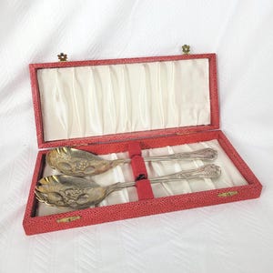 May include: A pair of silver-toned serving spoons with ornate embossed floral patterns on the bowls and handles, presented in a rectangular red textured case with a cream-colored satin interior lining and two red fabric loops to secure the utensils.